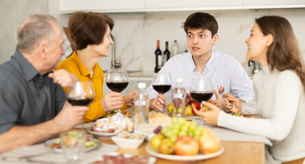 Young grown son with wife and parents having cheerful conversation at family holiday table in light kitchen