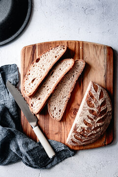 An artisanal loaf of sourdough bread with a decorative leaf pattern, sliced and displayed on a wooden cutting board alongside a serrated knife and a gray linen cloth.