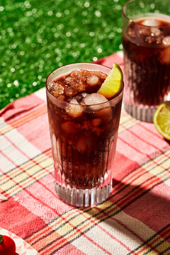 Glass of soda on picnic blanket on grass with sunlight, cherry tomatoes and lime, summer setting