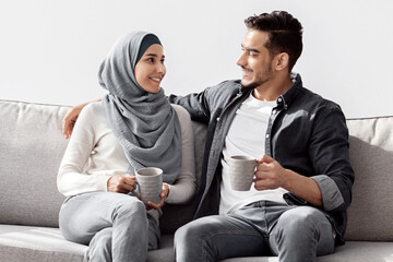 Loving middle-eastern young family husband and wife in hijab sitting on couch in empty room, drinking coffee while unpacking stuff, having conversation and smiling, planning house renovation