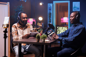 Cheerful black male talk show host engaging with his guest and online audience on live broadcast. African American men with wireless headset, sitting in home studio recording a vlog for social media.