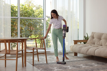 Smiling young woman cleaning rug with cordless vacuum cleaner at home