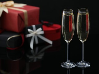 Delicious champagne in glasses and Christmas presents on black mirror surface, closeup. Space for text