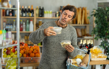 Focused man with shopping basket trying to make choice holding two sorts of herring in grocery...