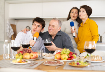 Joyful elderly man and woman sharing microphones with adult children and singing passionately during festive home gathering with food and drinks