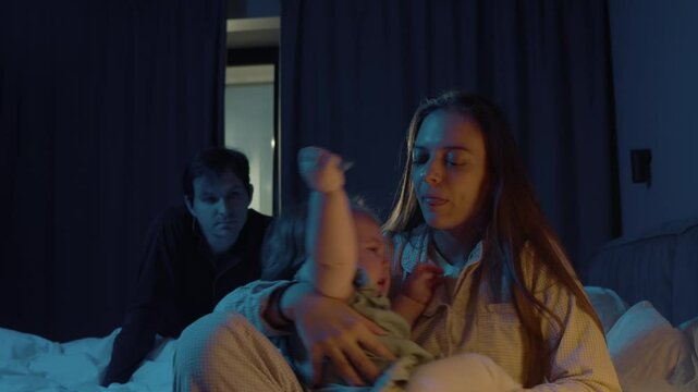 Young mother tries to comfort crying child, who has just woken up. She holds him in her arms, sitting on bed in their apartment in night. Disgruntled father looks on, sleepless.