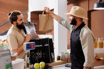 Delivery man is given a paper bag by male vendor. Caucasian courier picks up orders at the neighborhood convenience store and delivers natural, eco-friendly products to customers.