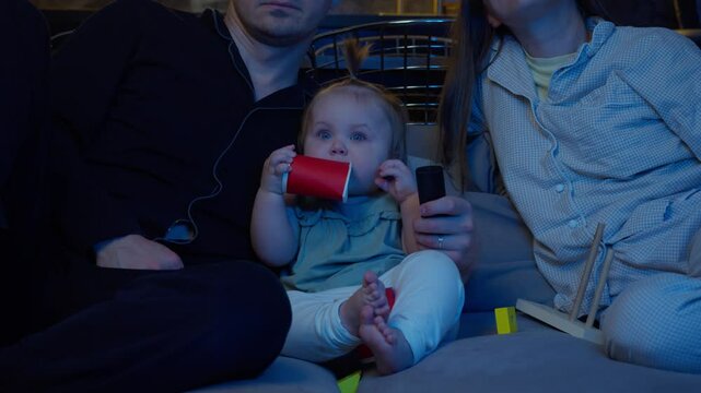 Cute toddler, about two year old, is watching cartoons on TV with great enthusiasm. He is sitting next to his parents, holding and nibbling on cardboard cup.