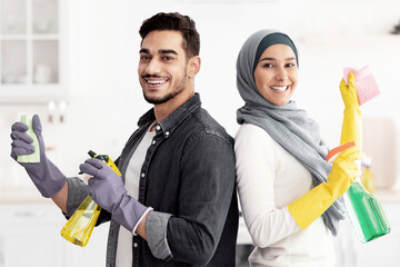 Cheerful middle-eastern handsome husband and pretty wife in hijab cleaning kitchen together,...