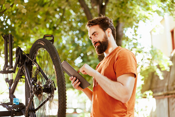 Healthy and active caucasian man multitasking inspecting and repairing his bicycle while using his digital tablet for guidance. Young sporty dedicated male cyclist surfing the internet to maintain