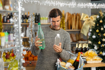 Portrait of focused glad positive man purchasing bottled water in small grocery store for christmas table