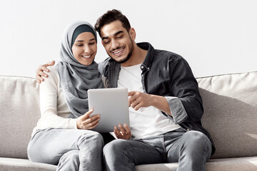 Joyful muslim family sitting on couch in new apartment, using pad, copy space. Happy middle-eastern young man and woman in hijab buying furniture for house, unpacking stuff after moving