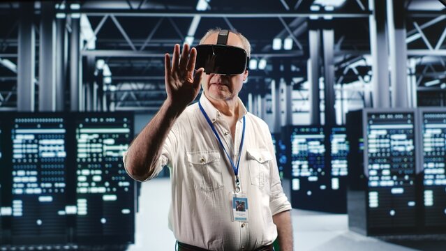 Elderly engineer in modern data center wearing virtual reality headset, performing server rigs maintenance. Experienced technician using vr glasses in high tech facility to do checkup on supercomputer