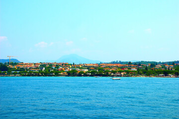 Hectic scenery with the town of Lazise, Lazise Hotel, the promenade with its lined green trees, a lot of pastel-colored buildings, hazy mountains in the background, cyan sky and rippled azure waters