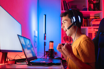 Gamer celebrating victory in an online multiplayer game match on a gaming laptop in his home room, illuminated by colorful LED lights, surrounded by streaming equipment and accessories © Marcio