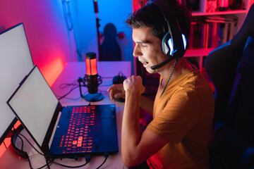 Gamer playing an online multiplayer game on a gaming laptop. He is in a dark room illuminated...
