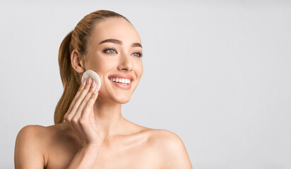 Facial Beauty Treatment. Happy Young Lady Using Cotton Pad Cleaning Face Looking Aside Over Gray Studio Background. Panorama, Free Space