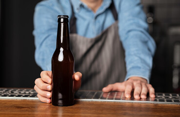 Barman in apron rests on wooden bar counter and serves bottle of beer without label, close up, cropped