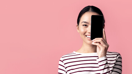 Fototapeta premium Cheerful Asian Female Covering Face With Blank Black Smartphone, Smiling Young Korean Woman Demonstrating Copy Space For Your Mobile Design And Advertisement, Pink Background, Mockup Image