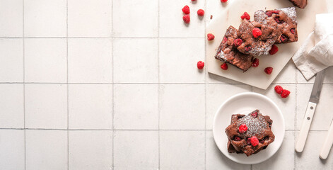 Board and plate with pieces of raspberry chocolate brownie on white tile background