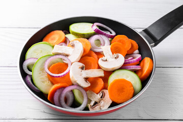 Frying pan with mix of fresh vegetables and mushrooms on white wooden table, closeup