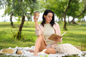 Pretty young woman with cup of coffee reading book on blanket in park