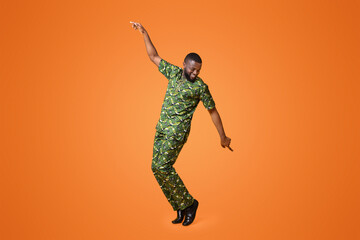 Carefree black guy in green african costume dancing over yellow studio background
