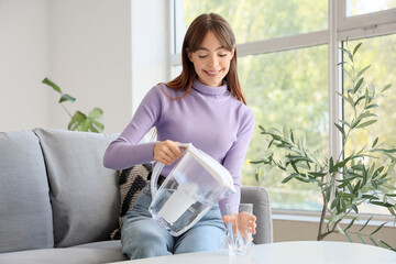 Young woman pouring water from filter jug into glass on table at home