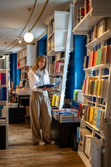 Enthusiastic woman selects books in bookstore alone. Female book lover browsing, searching...