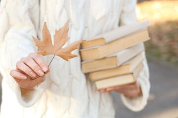 Woman holding stack of books and autumn leaf in park, closeup