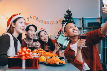 Group of young Asian man and women as friends having fun at a New Year's celebration, holding gift boxes standing by Christmas tree decoration, midnight countdown Party at home with holiday season.