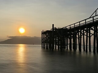 A weathered wooden pier extends into the water at sunrise, with barnacles clinging to the support...