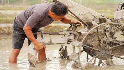 Indonesian adult male farmer installing a plough or plow onto a hand walking tractor on rice paddy field