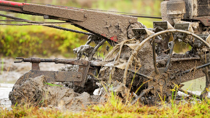 a close-up of a hand tractor's iron plow and wheels tilling soil in rice paddy field, farm tool for...