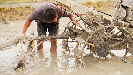 Southeast asian rice farmer installing a harrow farm tool on a hand tractor in muddy field for soil tilling