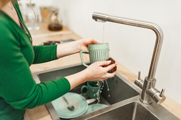 Cleaning, woman in green dress with green ceramic cup in kitchen for hygiene, routine and coffee mug. Female person, washing with water for disinfecting, protection and sink at home 