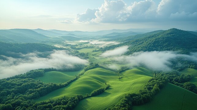 Aerial view of lush green fields and rolling hills with mist and mountains under a cloud-filled sky in a serene landscape scene.