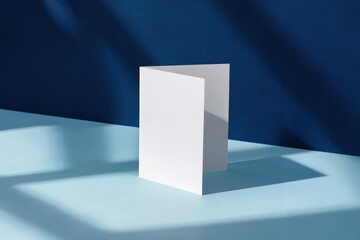 Fototapeta premium White blank folded card on light blue surface against deep blue background with soft lighting and gentle shadows
