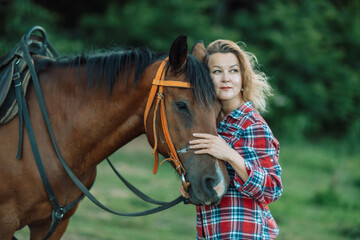 Obraz premium Happy blonde with horse in forest. Woman and a horse walking through the field during the day. Dressed in a plaid shirt and black leggings.