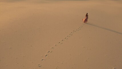 Aerial view from a drone flying next to a gorgeous woman in a pink dress walking on sand dunes in the desert. Traveling fashion girl cinematic footage. 4K Scenic nature