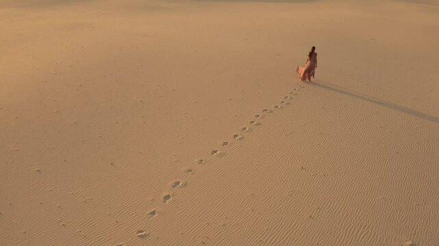 Aerial view from a drone flying next to a gorgeous woman in a pink dress walking on sand dunes in the desert. Traveling fashion girl cinematic footage. 4K Scenic nature