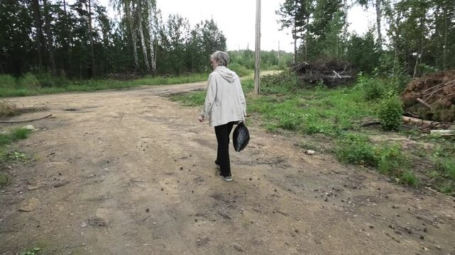 S2390029 Elderly woman in white jacket walking slowly and carrying package in right hand, coming to fork in road, rear view