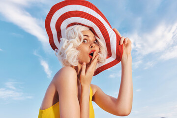 Surprised woman in a large striped hat with short blonde hair, yellow dress, against a blue sky...