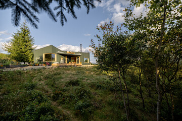 A green modern house sits on a gentle hill, surrounded by lush nature. Trees and greenery frame the scene, casting shadows in the evening light, highlighting a tranquil retreat amidst the forest.