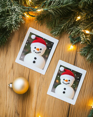 Christmas background with two polaroid photos of snowmen laying next to each other and Christmas tree branches with lights and small gold balls.