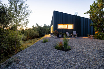 A modern cabin nestled in nature, illuminated warmly at dusk. Surrounded by lush greenery and trees, creating a serene escape in the wilderness