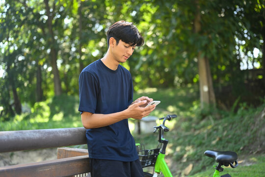 Young asian man dressed in casual clothes using smartphone near bicycle bike in the park