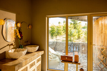A warm, sunlit bathroom featuring a double vanity with sculpted stone sinks, rattan cabinet...