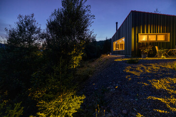 A modern cabin illuminated at twilight with a soft glow from interior lights. Surrounded by nature, a parked car and garden landscaping add to the serene and cozy atmosphere