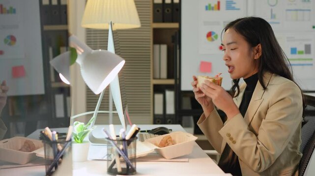 Asian businesswoman holding a sandwich while sitting in front of a computer during lunch break at her desk in a modern office.